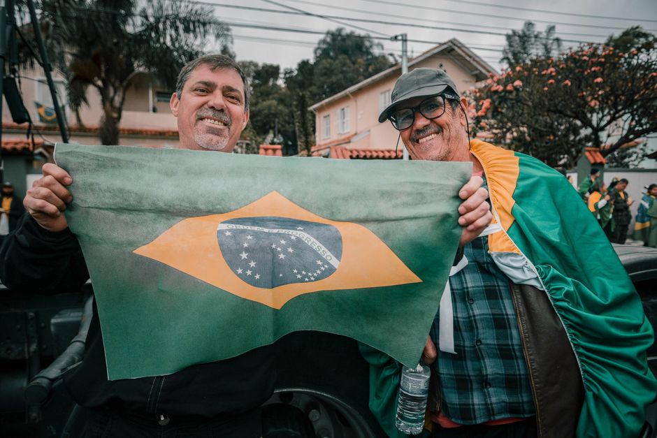 Two smiling men outdoors holding the Brazilian flag in a lively neighborhood.